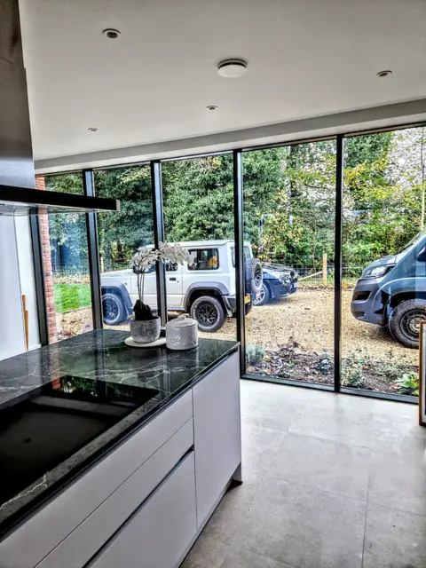 Full-height aluminium glazing seen from inside a kitchen, looking out to the drive